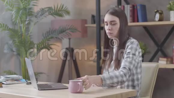 愤怒的白种人女孩从杯子里喝茶或咖啡开始在笔记本电脑键盘上打字令人厌烦的女性视频的预览图