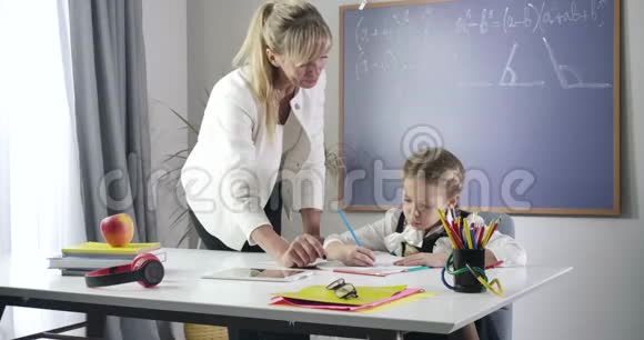 开朗的白种人女孩用红色铅笔在练习本上写字抬头看着成人老师可爱的女学生视频的预览图