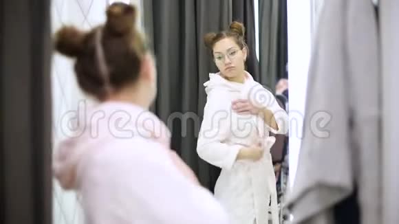 在服装店里穿浴袍的女孩在镜子里审视自己视频的预览图