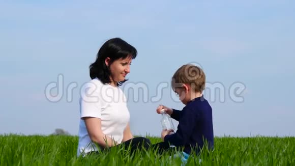 妈妈和儿子坐在绿草地上一个男孩在喝瓶子里的水视频的预览图