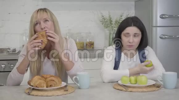 前面看到的悲伤丰满的高加索女人嫉妒地看着金发朋友吃美味的牛角面包和咀嚼苹果视频的预览图