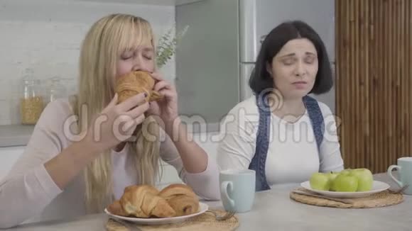 一幅令人沮丧丰满的高加索女人嫉妒地看着苗条的金发朋友吃美味的羊角面包和咬苹果的画像视频的预览图