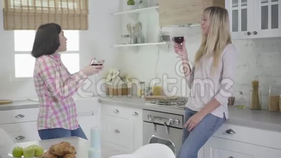 两个积极的白人妇女站在现代化的厨房里拿着酒杯聊天快乐的女性朋友视频的预览图