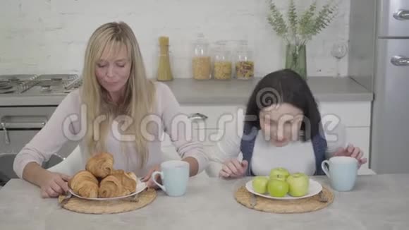 布鲁内特高加索女人闻着绿苹果她的金发朋友从盘子里拿出牛角面包健康和健康视频的预览图
