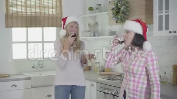 两个戴着红色圣诞帽的白种人成年女性在现代厨房跳舞唱歌喝着酒快乐的朋友视频的预览图