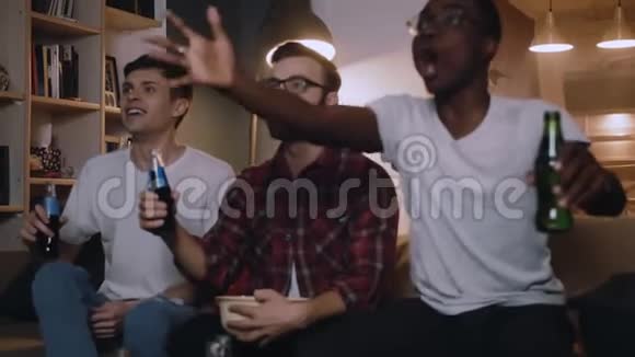 快乐的多民族年轻男性朋友在电视上看游戏享受饮料微笑和交谈享受娱乐慢动作视频的预览图