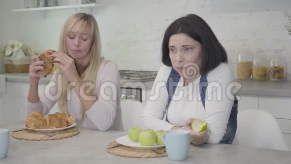 肥胖的白人女人在嚼苹果她苗条的朋友给她的盘子里放着美味的羊角面包快乐的女士在吃苹果视频的预览图
