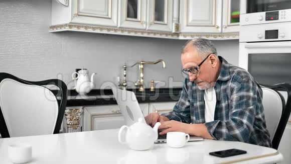 专注于家中老年商业的男性在家中厨房里用手提电脑喝茶视频的预览图