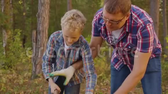 爸爸一起儿子在夏天的森林里用斧头砍柴父亲和儿子用斧头砍木头视频的预览图