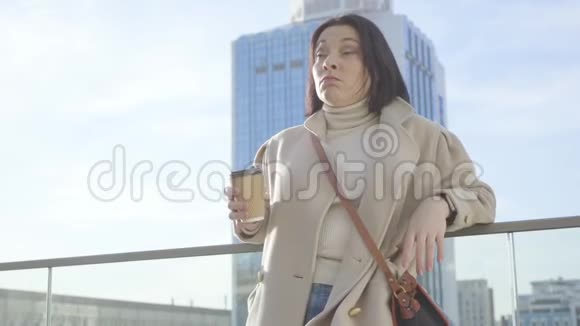 年轻的褐发白种人女人打哈欠和喝咖啡站在大建筑的背景肖像视频的预览图