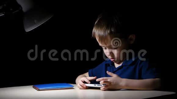 一个小男孩晚上坐在黑暗的房间里的桌子旁他的脸被桌灯照亮了一个快乐的孩子看着视频的预览图