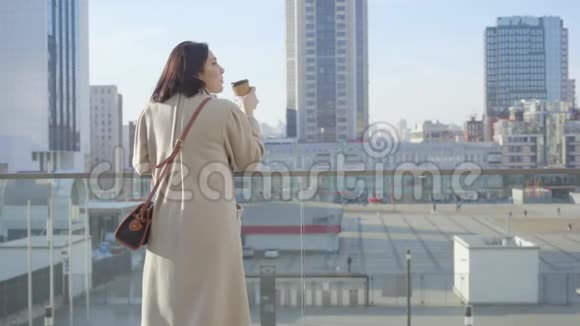 年轻的白种人女人喝咖啡和看城市街道的后景穿着优雅外套的成功女人视频的预览图