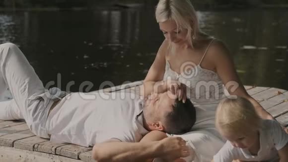 在一个阳光明媚的日子里年轻的家庭母亲父亲和儿子坐在河边的码头上男人躺在女人腿上视频的预览图