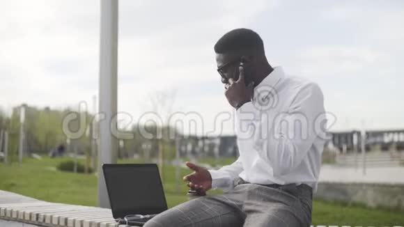 在城市公园边看带眼镜的非洲商人在电话和喝咖啡视频的预览图