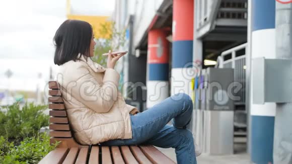 年轻的微笑的女人使用智能手机坐在购物中心附近的长凳上美丽的欧洲女孩做语音留言视频的预览图