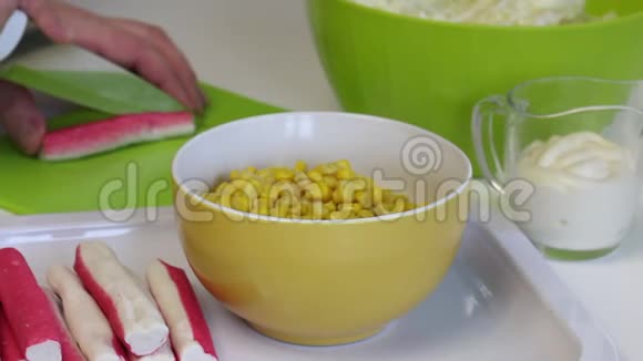 男人做沙拉切片蟹棒在切割板上前景是一个容器与玉米和蛋黄酱特写视频的预览图