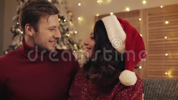 亲密的年轻美丽的爱情情侣微笑着看着彼此坐在靠近圣诞树的沙发上视频的预览图