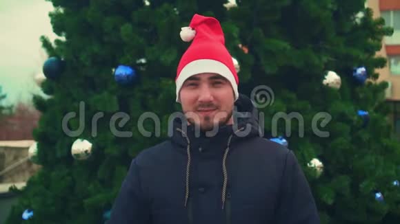 有胡子的年轻人戴着圣诞帽背景是圣诞树快乐的男人用他的手显示出好的标志视频的预览图