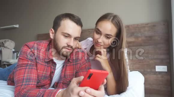 年轻的一家人躺在卧室的床上看着智能手机上的在线新闻视频的预览图