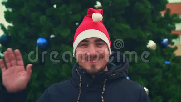 有胡子的年轻人戴着圣诞帽背景是圣诞树快乐的人挥手作为问候的标志视频的预览图