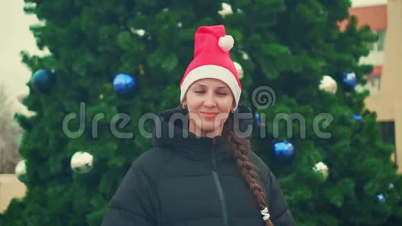 圣诞树背景上戴着圣诞帽的年轻女子幸福漂亮的女人微笑着用一只超级手示意视频的预览图