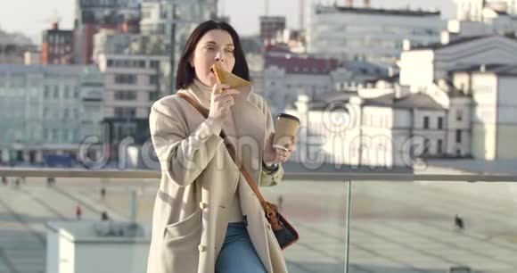 高加索年轻女子站在城市街道上端着咖啡杯吃着三明治的画像自信的黑发女人视频的预览图