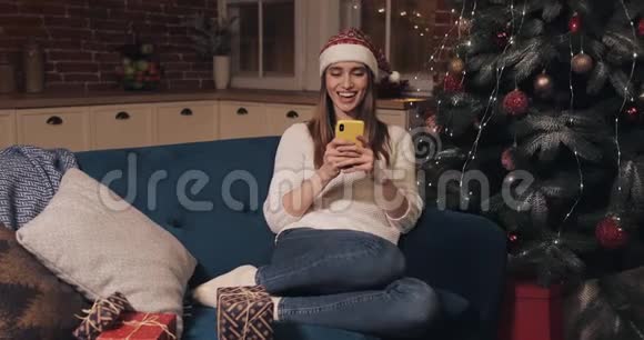 戴着圣诞帽的白种人金发女孩在圣诞家庭背景下快乐地坐在沙发上视频的预览图