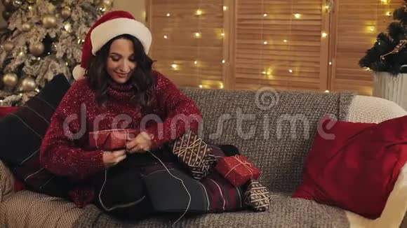 圣诞老人帽里美丽的年轻女性在沙发上坐着的碗里完成了圣诞礼物绑带和绑带视频的预览图