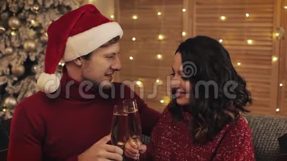 年轻夫妇坐在沙发上靠近圣诞树在舒适的家背景下闪烁着香槟的眼镜可爱的家庭视频的预览图