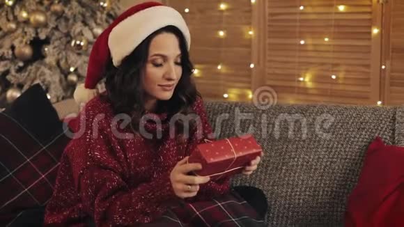圣诞帽里年轻漂亮的布鲁内特女士坐在灰沙发的圣诞树旁手里拿着红色的礼物盒视频的预览图