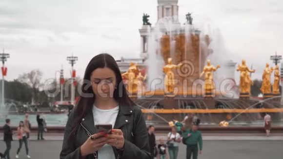 漂亮的女孩穿着黑色夹克和白色t恤在智能手机上输入短信关门视频的预览图