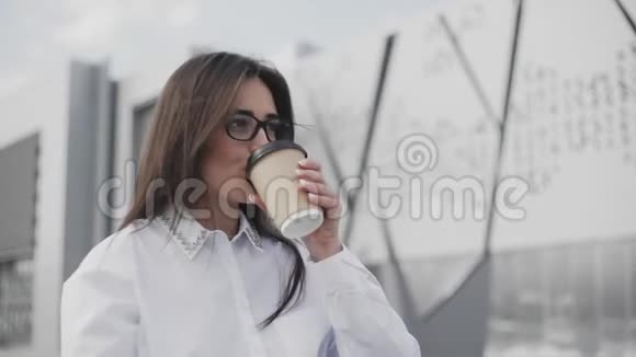 漂亮的女商人戴着眼镜坐在办公室附近的公园里一边说着智能手机一边喝着咖啡视频的预览图