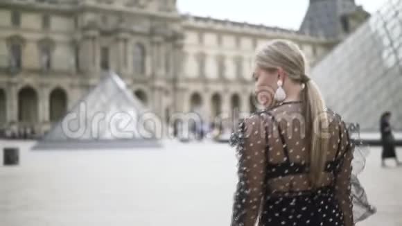 一个阳光明媚的夏日美丽的金发女人穿着透明的点缀裙走在卢浮宫博物馆前行动视频的预览图