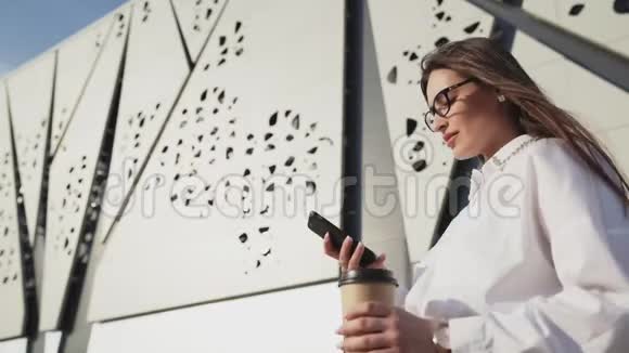 漂亮的商务女性戴着眼镜看着智能手机喝着咖啡现代背景建设视频的预览图