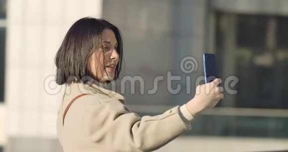 开朗的高加索女人自拍的肖像年轻的黑发女士用智能手机在城市大街拍照视频的预览图