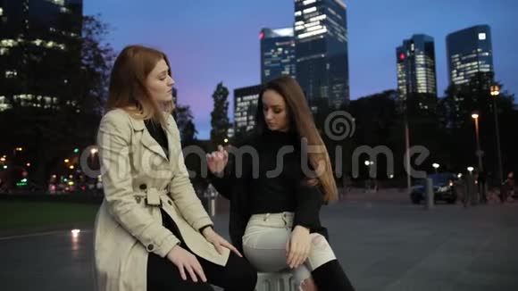 两个年轻漂亮的女孩晚上坐在城市公园的手提箱里等着视频的预览图