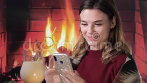 漂亮的微笑女孩坐在壁炉背景下的艾格诺格圣诞鸡尾酒旁手里拿着电话视频的预览图