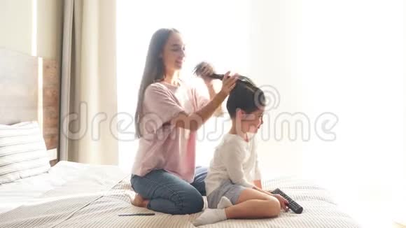 年轻的母亲穿着随便坐在大床上梳着小黑发女儿的卷发视频的预览图
