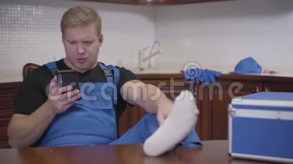 坐在厨房里用智能手机的懒惰军人的肖像穿着蓝色工作服的白种人成年男子视频的预览图