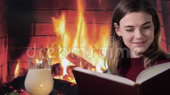 年轻女子在壁炉旁看一本有趣的书喝一杯蛋酒的圣诞鸡尾酒的画像视频的预览图