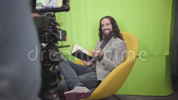一位英俊的中东男人坐在黄色的扶手椅上拿着书在镜头前交谈摄像机录音视频的预览图