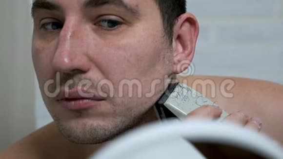 一个男人在家里用电动剃须刀刮胡子和胡子在镜子前脱毛男人照顾视频的预览图