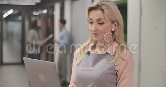 一位金发白种人成年女性的特写镜头她拿着笔记本电脑站在开放空间的办公室里看着摄像机微笑着视频的预览图
