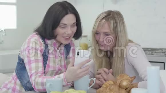 特写两名白人成年女性看着智能手机屏幕微笑积极的女性朋友在室内休息视频的预览图