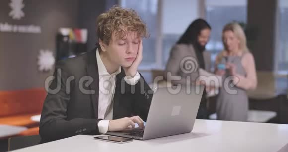 疲惫的白人年轻人在笔记本电脑键盘上打字看着智能手机揉着头的肖像精疲力竭的男性视频的预览图