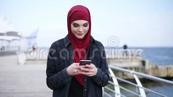 迷人的年轻女孩头上戴着头巾一边微笑着给某人发短信一边在她身上滚动一些东西视频的预览图
