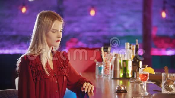 漂亮的金发女郎涂着红色口红坐在酒保摊边喝着吸管里的红色饮料视频的预览图
