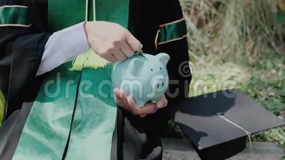 穿着学士制服的人正在把硬币放进蓝色储蓄罐为教育计划和退休基金存钱视频的预览图