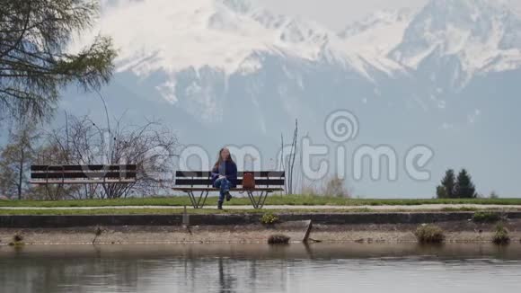 迷人的女孩坐在一张木凳上在湖和山上有着惊人的景色享受风景如画的女人视频的预览图