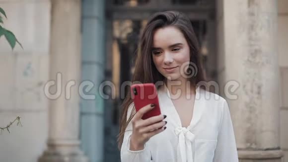 美丽的微笑布鲁内特女孩走在科西老城建筑背景同时使用她的手机在红色的情况和视频的预览图